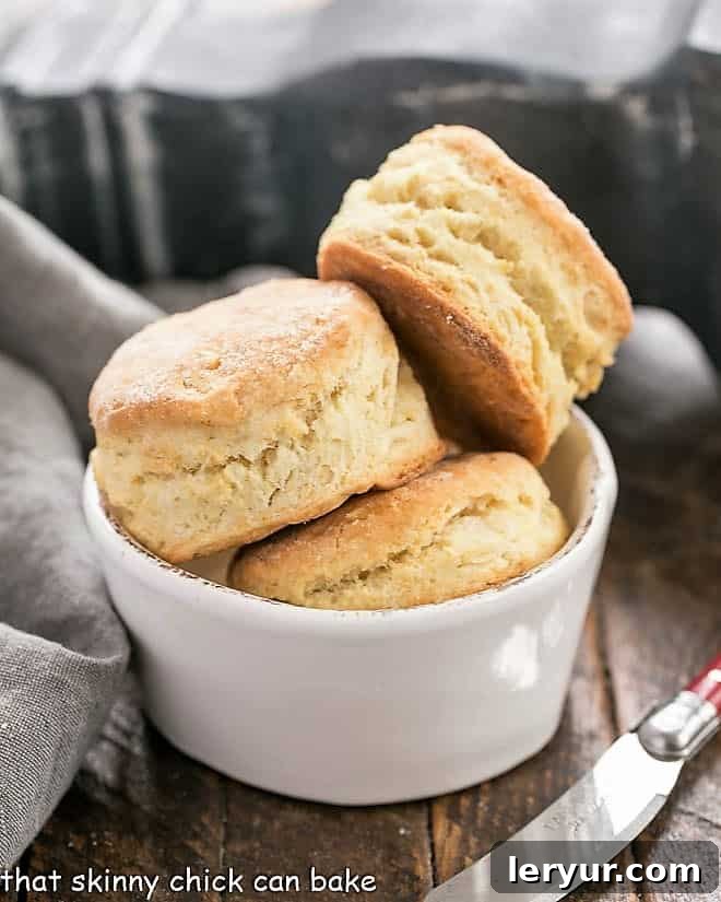 A pristine white bowl brimming with freshly baked, golden-brown homemade buttermilk biscuits. The image beautifully captures their inviting texture and generous size, hinting at their deliciousness and readiness to be savored.