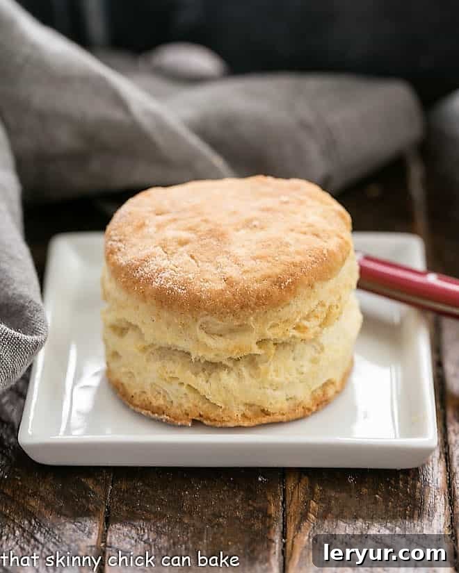 A golden-brown buttermilk biscuit on a square white plate, accompanied by a red-handled knife, signifying its readiness to be enjoyed.