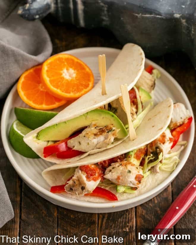 Two perfectly grilled fish tacos generously filled with fresh shredded cabbage, creamy avocado, and succulent white fish, all drizzled with a zesty sauce and presented on a pristine white plate, ready to be enjoyed.