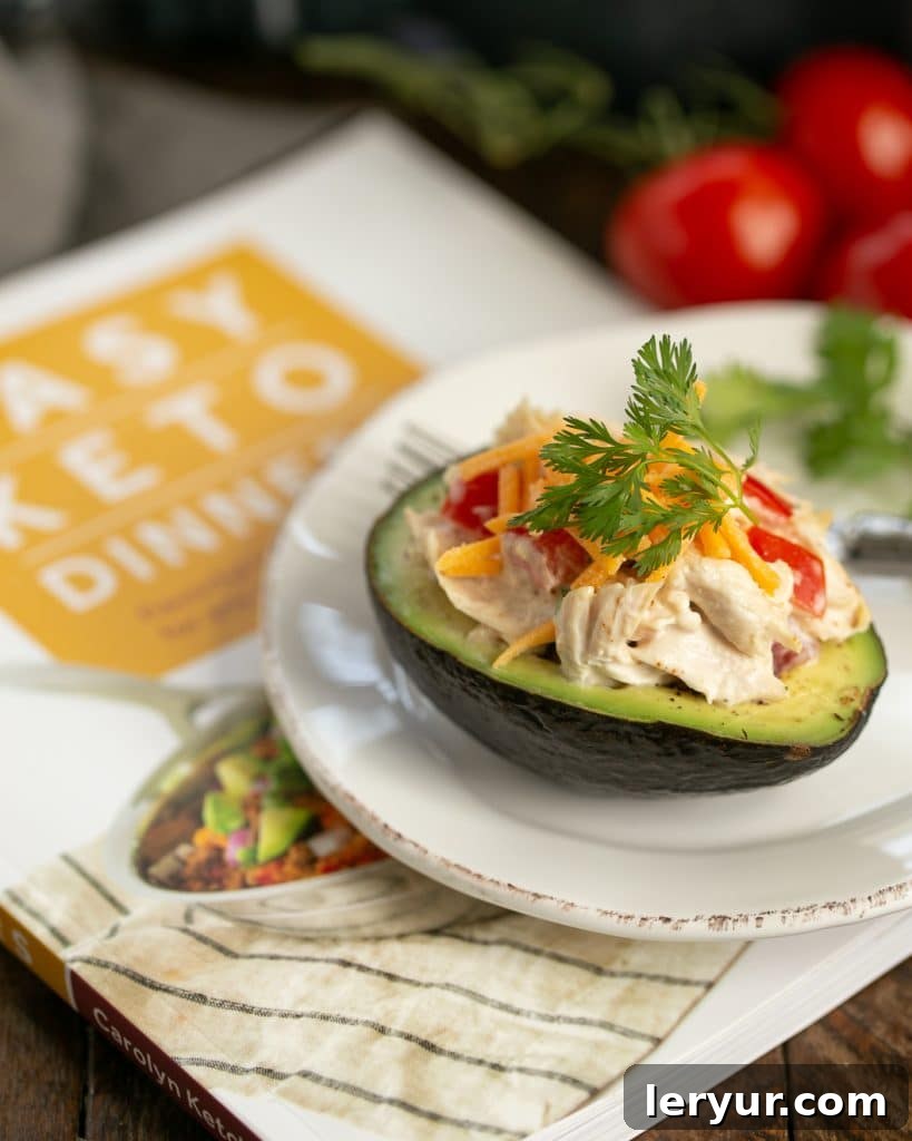 A single Southwestern Chicken Stuffed Avocado half, garnished with a sprinkle of cheddar cheese, is presented on a white plate, resting elegantly on Carolyn Ketchum's 'Easy Keto Dinners' cookbook. This image highlights the recipe's origin and delicious appeal.