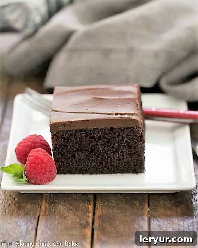 Black Magic Snack Cake - an old fashioned chocolate buttermilk cake with chocolate buttercream A slice of Black Magic Snack Cake on a white square plate with a raspberry garnish, showcasing its moist texture and rich chocolate frosting.