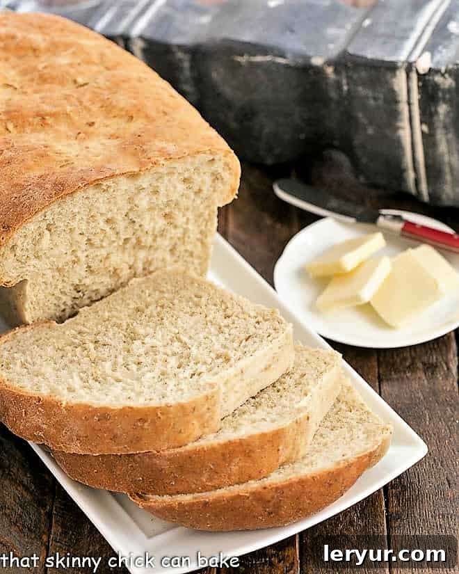 Oat Bread Recipe - a sweet, hearty bread flavored with a touch of brown sugar A partially sliced loaf of warm oatmeal bread on a white serving tray, with a plate of butter pats ready in the background.