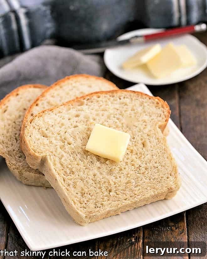 Three golden-brown slices of classic oatmeal bread on a square white plate, with a dollop of butter melting on the top slice.
