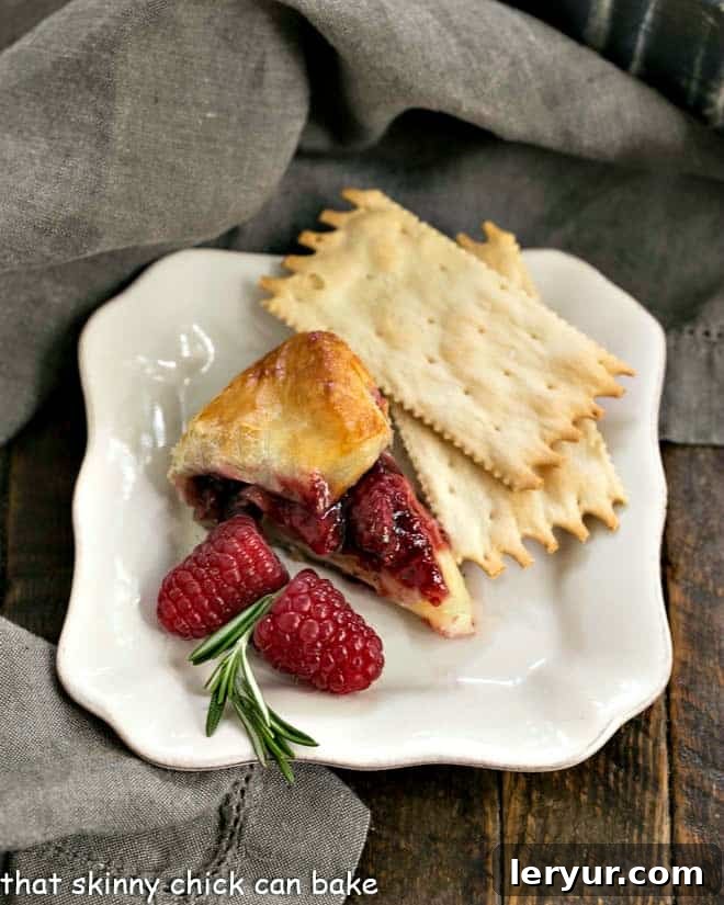 Baked Brie with Raspberries - a spectacular appetizer with creamy brie, a raspberry, rosemary topping and a flaky puff pastry crust! Slice of raspberry brie en croute on an appetizer plate with crackers and fresh raspberries, ready to serve.