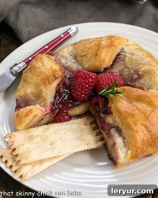 Raspberry Brie en Croute Baked - a spectacular appetizer with creamy brie, a raspberry, rosemary topping and a flaky puff pastry crust! Raspberry Brie en Croute on a serving plate with crackers and a red handled knife, ready for guests.
