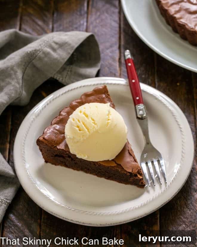 Chocolate Brownie Tart - a fudgy brownie served in wedges with ice cream and chocolate sauce! A slice of brownie tart topped with a scoop of vanilla ice cream on a round white plate with a red handled fork.