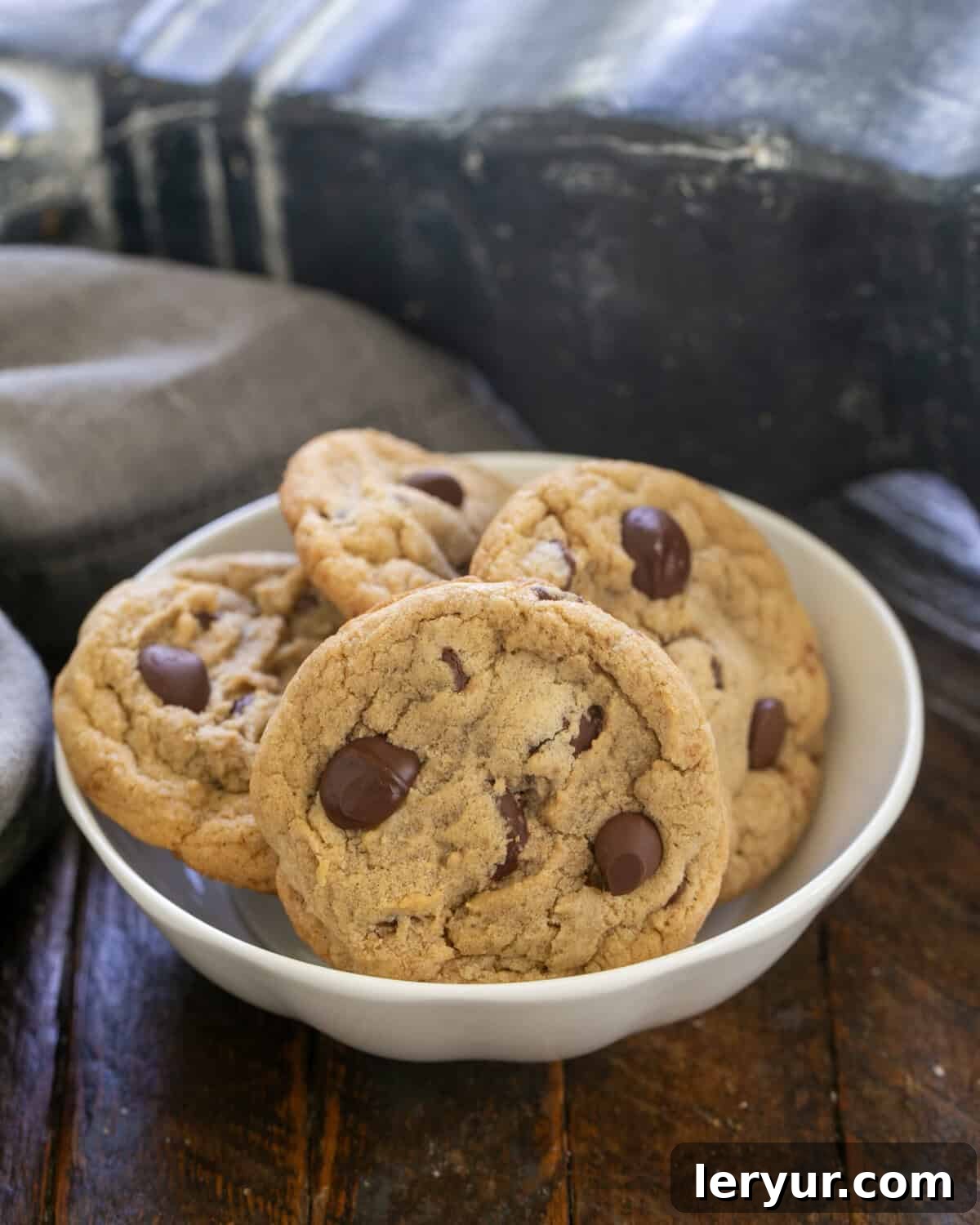 Killer Chocolate Chip Cookies made with dark brown sugar, two kinds of chocolate chips and an extra yolk for tasty, chewy results! Four perfectly baked, golden-brown killer chocolate chip cookies adorned with melty chocolate chips, artfully arranged in a white ceramic bowl, invitingly presented for indulgence.