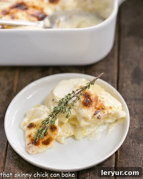 French Potato Gratin - A exquisite creamy cheesy potato casserole kissed with garlic, thyme and topped with Gruyere cheese! The perfect holiday side. A serving of French Potato Gratin on a small white plate, garnished with a sprig of fresh thyme, showcasing its golden-brown crust and creamy interior.