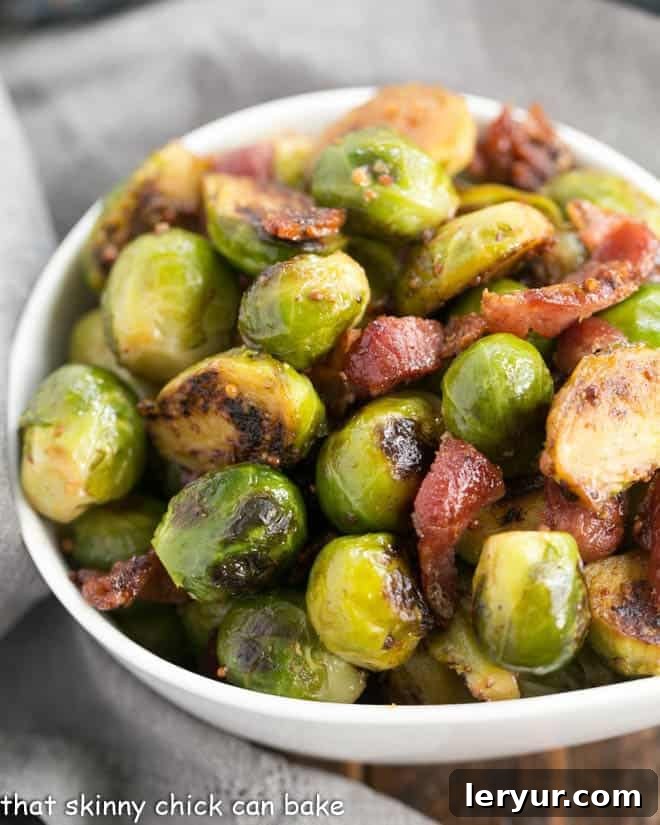 Maple Syrup Mustard Glazed Brussels Sprouts - an irresistible autumnal side dish with steamed, then sauteed Brussels sprouts glazed with the bold flavors of grainy mustard, garlic, shallots, real maple syrup and bacon! Maple Syrup Mustard Glazed Brussels Sprouts closeup in a white ceramic bowl, showcasing the glaze and bacon bits.
