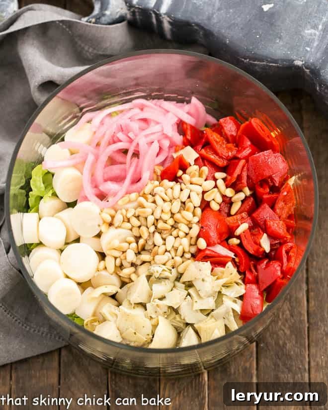 All the fresh ingredients for Italian Chopped Salad, including crisp lettuces, peppers, artichoke hearts, and hearts of palm, neatly arranged in a clear glass salad bowl before mixing.