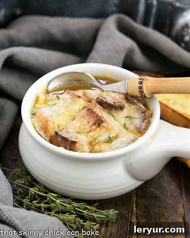 Classic French Onion Soup - A rich onion laden broth flavored with herbs, white wine and cognac and topped with cheesy toast! Classic French Onion Soup in a white soup bowl with a bamboo handled spoon, showcasing the rich broth and cheesy crouton.