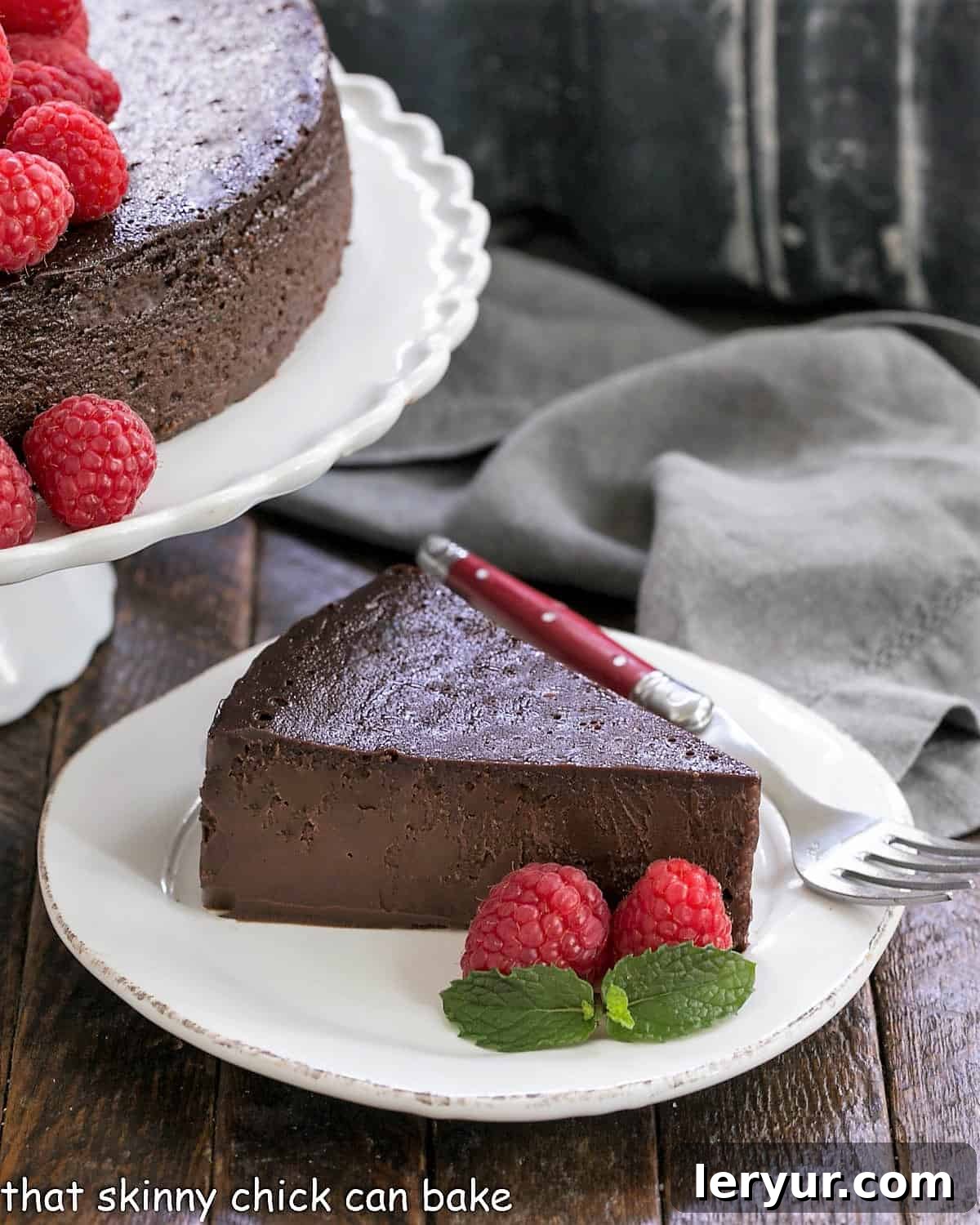 Flourless Chocolate Kahlua Cake - a rich, dense chocolate cake enhanced by coffee liqueur A decadent slice of Flourless Chocolate Kahlua Cake with fresh raspberries and a mint sprig, next to the full cake on an elegant cake stand.