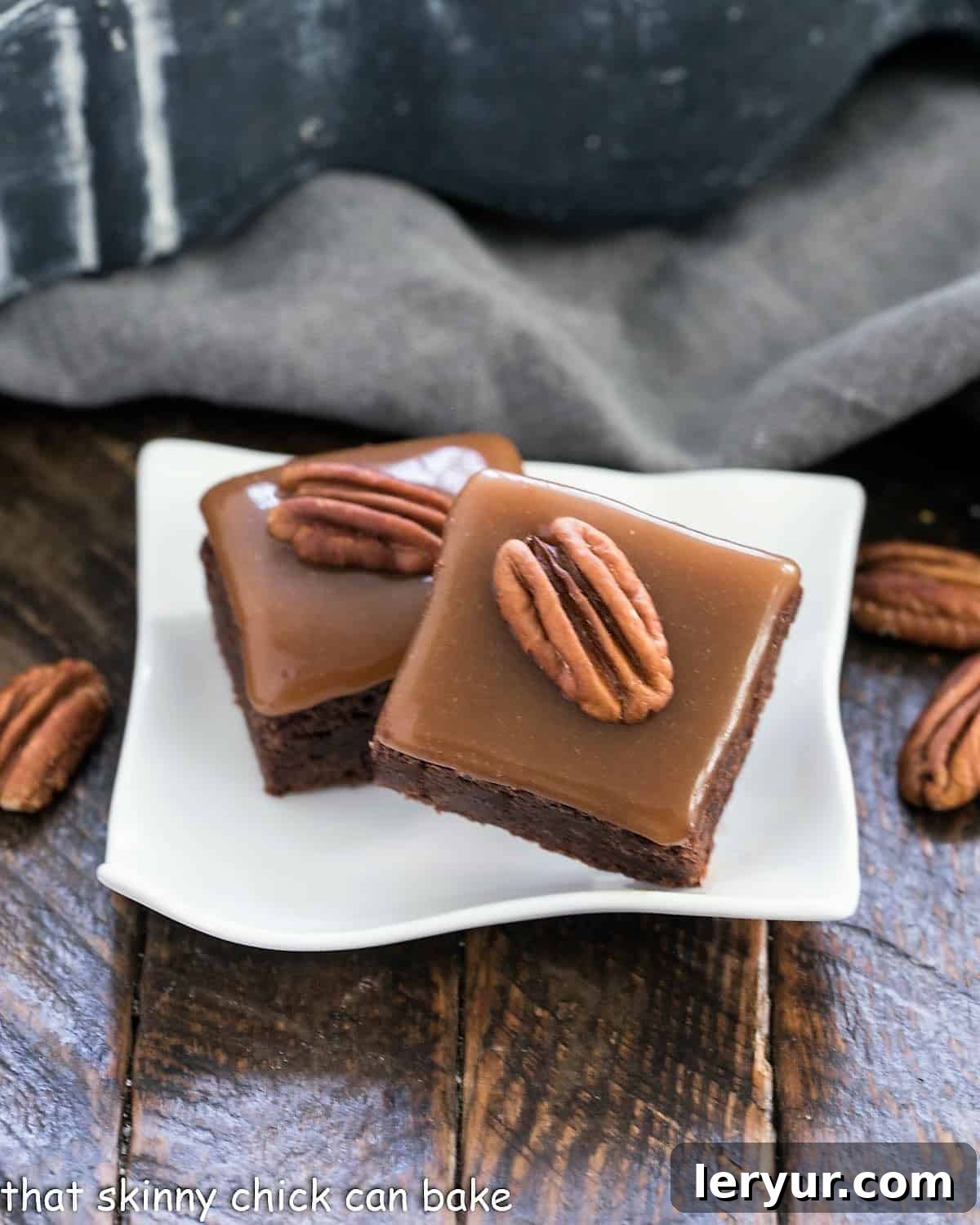 The Ultimate Turtle Brownies - fudgy brownies topped with the perfect chewy caramel and pecan halves Two decadently layered turtle brownies showcasing their fudgy chocolate base, gooey caramel, and pecan topping, resting on a small white wavy plate.