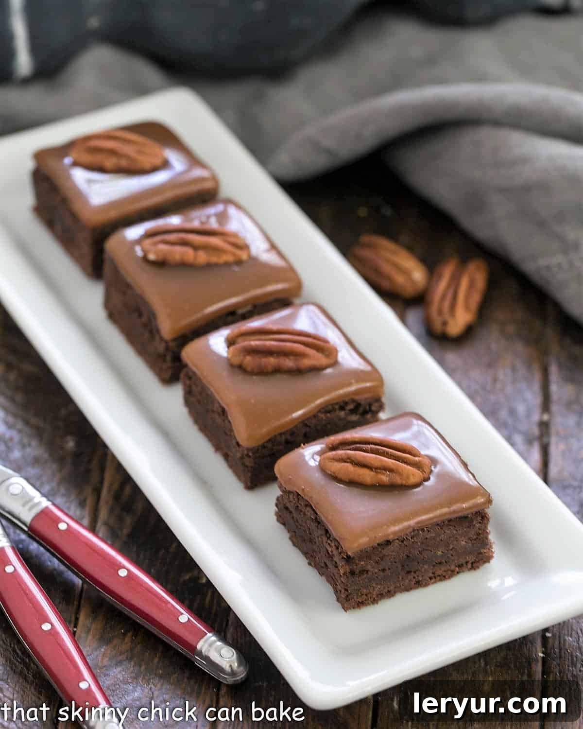 The Ultimate Turtle Brownies - fudgy brownies topped with the perfect chewy caramel and pecan halves A white serving tray laden with perfectly arranged caramel brownies, each adorned with a pecan half, accompanied by two elegant red-handled forks, ready for serving.