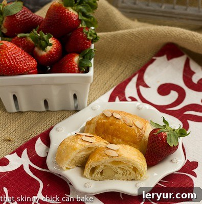 Almond croissants on a white plate on a red and white napkin with a bowl of fresh strawberries. Garnished with sliced almonds.