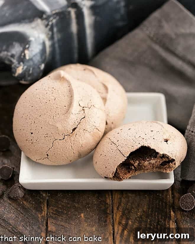 Three enticing chocolate meringues, two whole and one with a bite taken out, revealing its perfectly chewy, chocolatey center. They sit on a light background, highlighting their rich color and inviting texture.