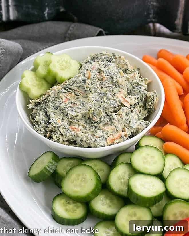Delectable Sour Cream Spinach Dip - a simple yet elegant party dip recipe, much tastier than any store-bought alternative, ideal for showcasing fresh produce. A white ceramic bowl filled with creamy Sour Cream Spinach Dip, perfectly chilled and ready to serve, sits on a rustic white crudité platter surrounded by fresh vegetables.