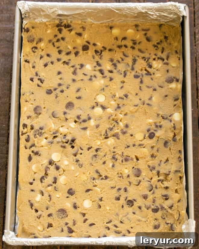 Spreading the dense, chocolate chip-filled cookie dough evenly into a baking pan lined with foil or parchment paper, preparing it for the oven.