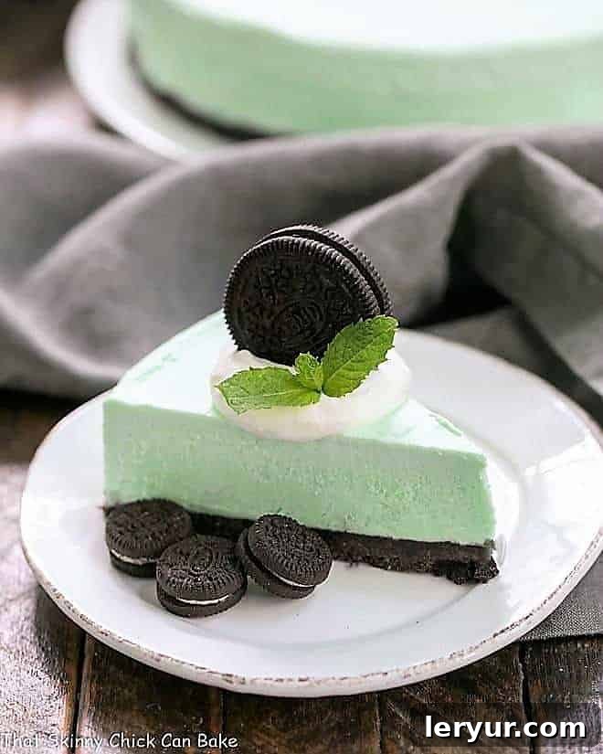 Frozen Grasshopper Pie with an Oreo Crust - Mint lovers will swoon with every bite of this frozen, no bake dessert! A slice of Frozen Grasshopper Pie with an Oreo Crust on a small round white plate, topped with whipped cream and an Oreo cookie, showcasing its vibrant green filling.