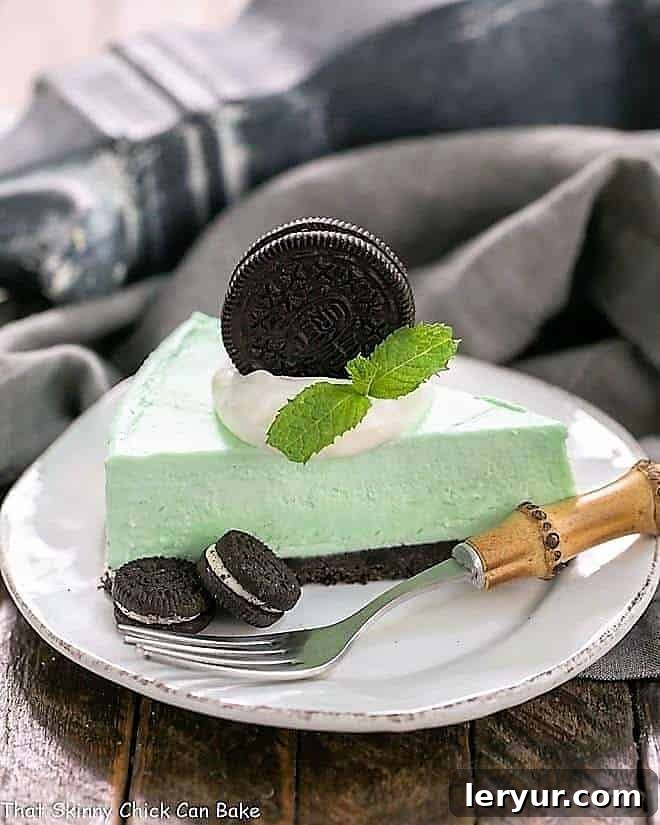 Minty, green, frozen No-Bake Dessert with an Oreo Crust - Mint lovers will swoon with every bite of this frozen no bake dessert! A perfect slice of Frozen Grasshopper Pie with an Oreo Crust on a white dessert plate, accompanied by whole Oreo cookies and a bamboo-handled fork, highlighting its inviting texture.