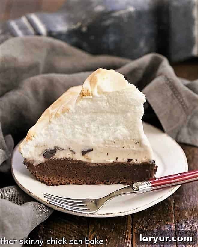 A perfectly portioned slice of Brownie Baked Alaska on a white dessert plate, revealing distinct layers of fudgy brownie, creamy vanilla ice cream with chocolate chips, and a golden toasted meringue.