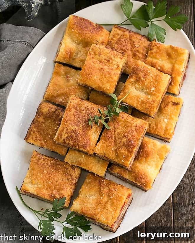 An overhead shot showcasing freshly baked Antipasto Squares on a white oval platter, garnished with a sprinkle of fresh herbs.
