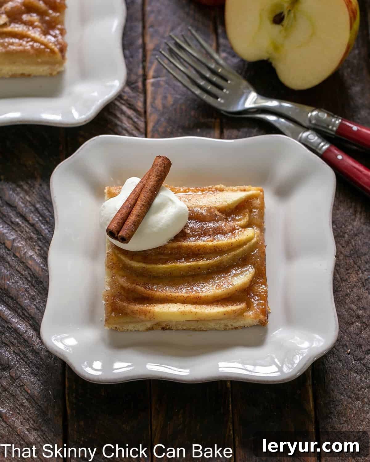 slice of apple kuchen on a square white plate with whipped cream and a cinnamon stick garnish.