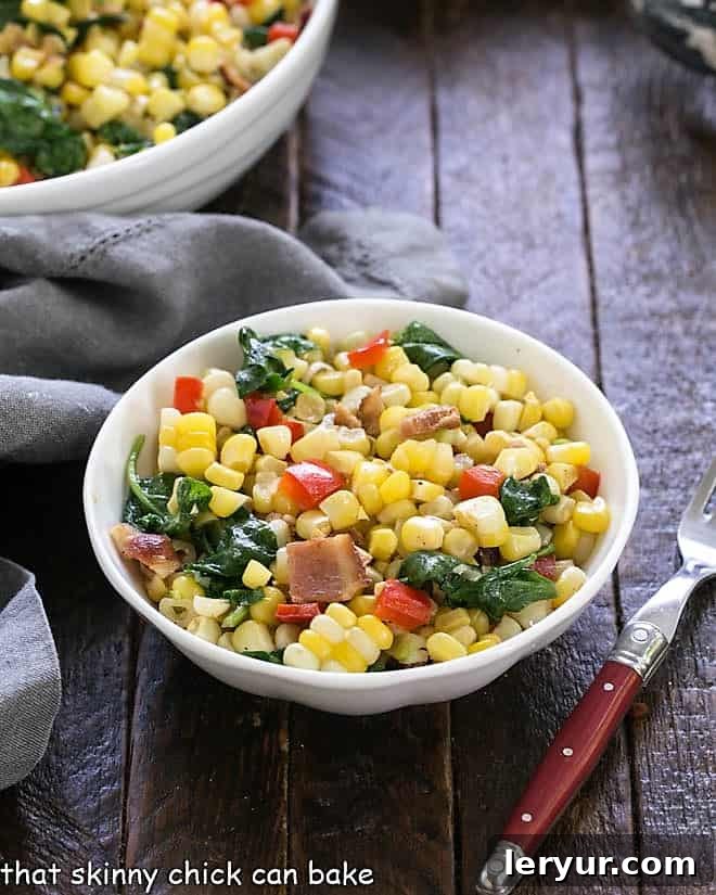 Skillet corn with spinach and peppers in a small white bowl, ready to be served with a red handle fork.