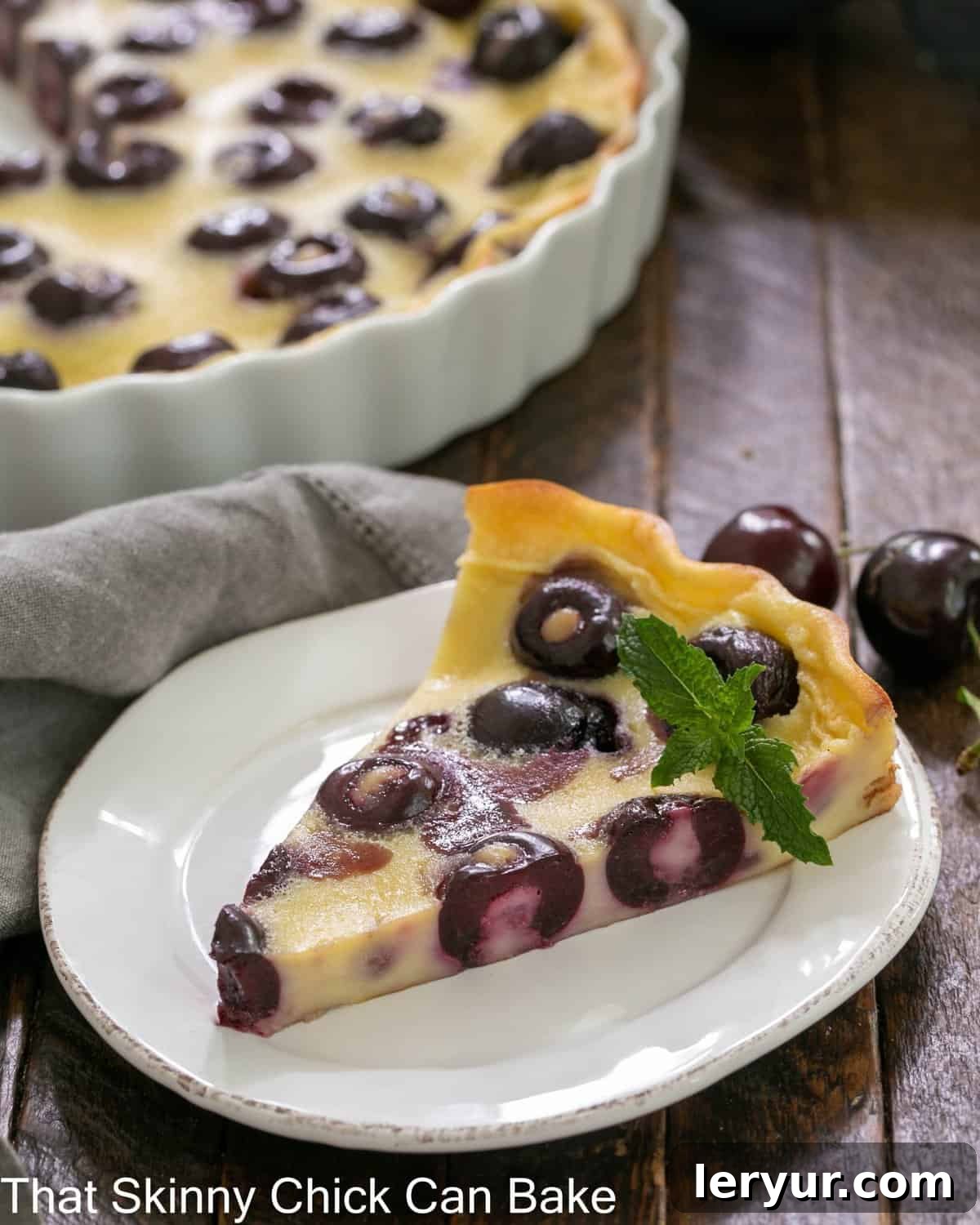 Rustic Whole Cherry Clafoutis 2 A perfectly sliced piece of cherry clafoutis on a white plate, showcasing the juicy cherries and golden-brown crust. A white ceramic baking dish is visible in the background.
