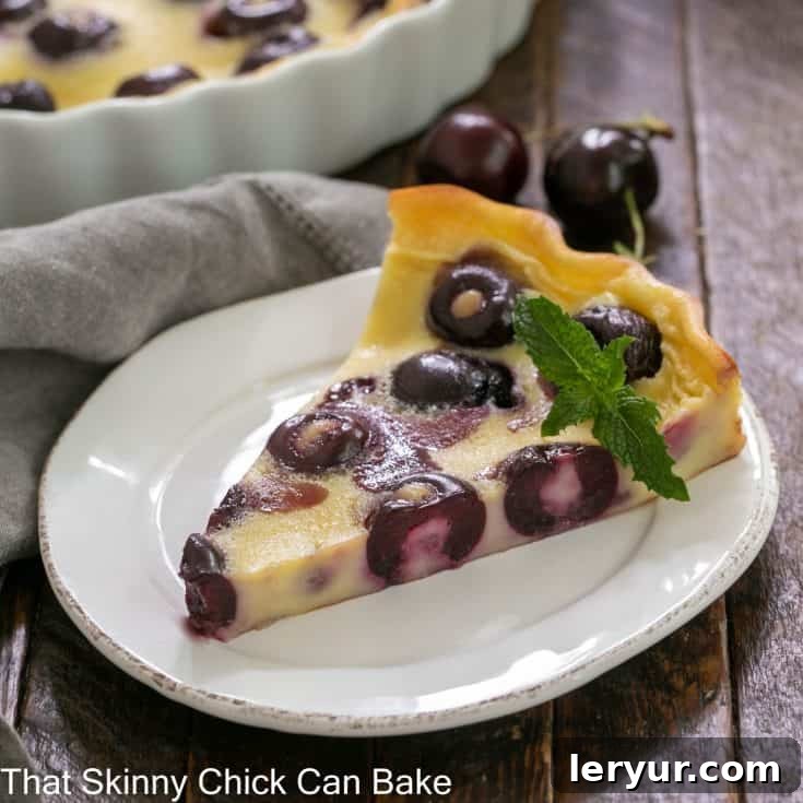 A beautifully presented slice of cherry clafoutis on a white plate, garnished with a sprig of fresh mint. The texture shows the custard and embedded cherries.