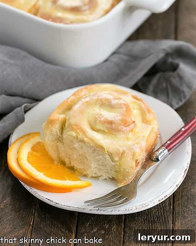A beautiful array of homemade orange sweet rolls in a baking dish, with one individual roll placed on a small white plate beside a red-handled fork, inviting indulgence.