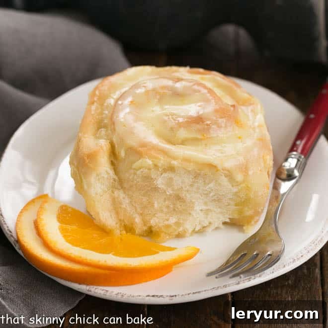A single homemade orange roll on a round white plate, garnished with fresh orange slices, next to a red-handled fork.