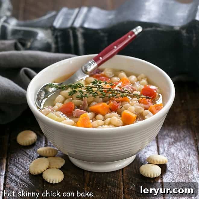 Bowl of bean soup with ham garnished with thyme in a white bowl with a red handle spoon surrounded by oyster crackers