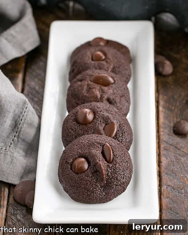 A narrow white ceramic tray holds a line of freshly baked chocolate chocolate chip cookies, showcasing their rich color and inviting texture.