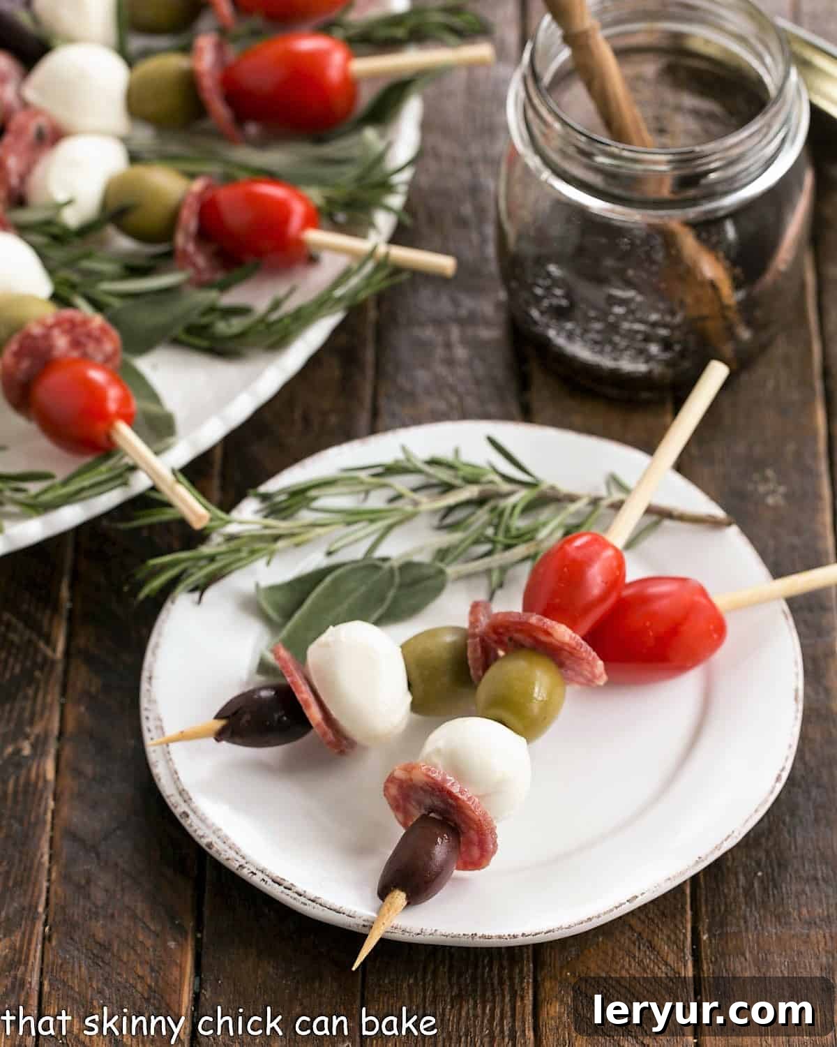 Two perfectly threaded antipasto skewers resting on a white appetizer plate, accompanied by a fresh rosemary garnish.