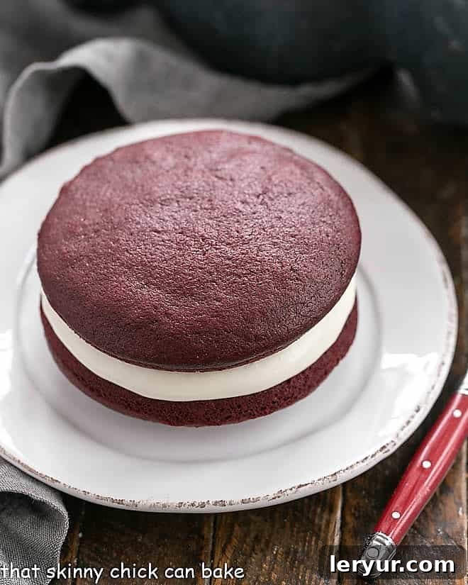 Red Velvet Whoopie Pie Recipe: A delightful single serving of this cake-like sandwich cookie. A single red velvet whoopie pie on a round white plate, showing the creamy filling peeking out.