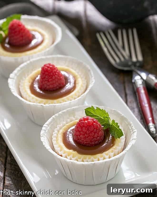 Caramel Topped Mini Cheesecakes on a white rectangular tray garnished with raspberries and fresh mint leaves, showcasing their golden caramel drizzle and vibrant fruit.