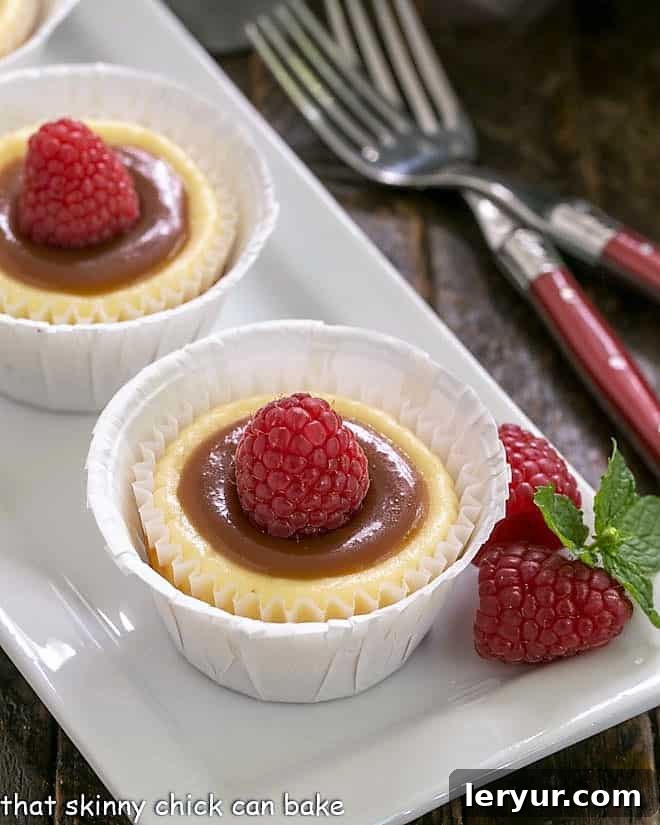 Two Caramel Topped Mini Cheesecakes, elegantly drizzled with caramel sauce and garnished with a single raspberry on a pristine white tray.