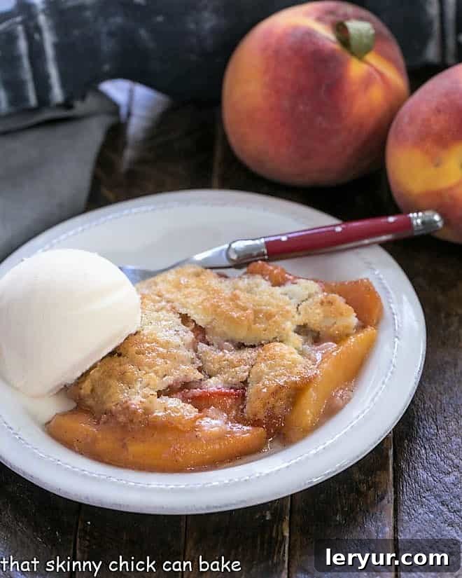 A delightful serving of easy peach cobbler, crowned with a scoop of melting vanilla ice cream, presented on a white plate with two fresh, ripe peaches in the soft background.